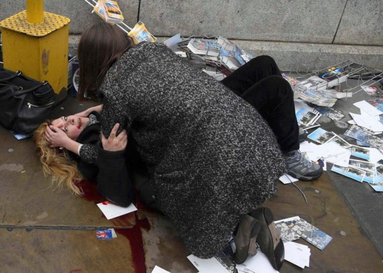 Photograph of Woman injured in Westminster Bridge terrorist vehicle attack, March 2017, laying on the ground bleeding while another woman leans over her trying to apply direct pressure to a wound on her head. Blood trails on the ground running away from her.