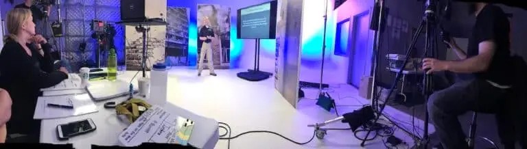 A professional film stage showing Dr. Shertz standing in front of high risk environment stage pieces and a television projecting slides while the crew watches with rapt attention