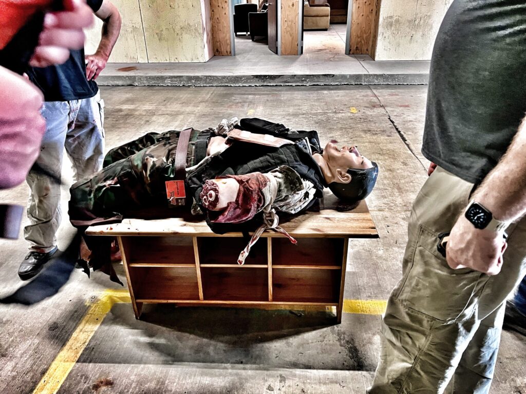 Using improvised equipment as litters - Frank the manikin with multiple amputations lays on a light weight coffee table rescuers battlefield recovered to carry him on.