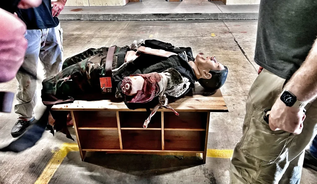 Using improvised equipment as litters - Frank the manikin with multiple amputations lays on a light weight coffee table rescuers battlefield recovered to carry him on.