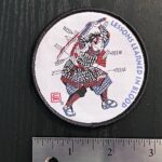 A woven patch with marrow edge and velcrow backing showing a Samurai in battle, bloodied and shot full of arrows, with a broken and bloodied sword raised overhead