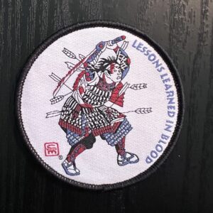 A woven patch with marrow edge and velcro backing showing a Samurai in battle, bloodied and shot full of arrows, with a broken and bloodied sword raised overhead