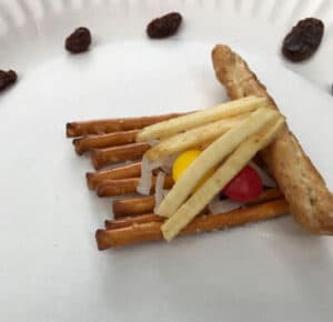 A fire model made by small kids with pretzels, matchstick potatoes, skittles, and coconut
