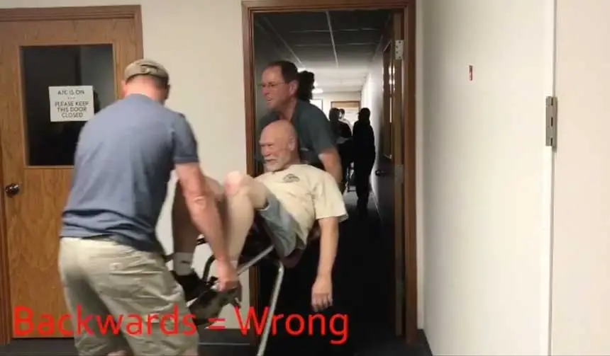 Two rescuers demonstrate bad carrying technique by walking backwards (they do get bonus points for using a chair to carry the casualty between them).