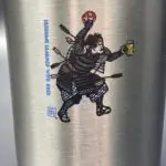 A closeup of the Samurai logo, festooned for Oktoberfest in Bavarian Blue & White, holding a pretzel overhead and a beer in his left hand